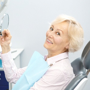 An older woman holding a small mirror and smiling after learning she is eligible for dental implant surgery