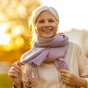 An older woman wearing a scarf stands outside and smiles after learning how affordable dental implants can be